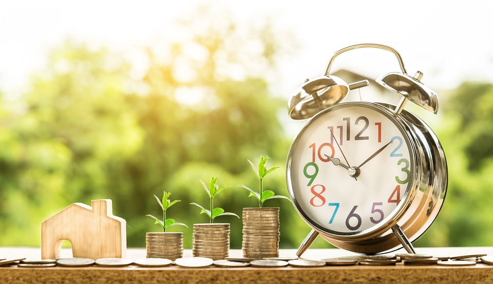 A wooden toy house next to a stack of coins and an alarm clock