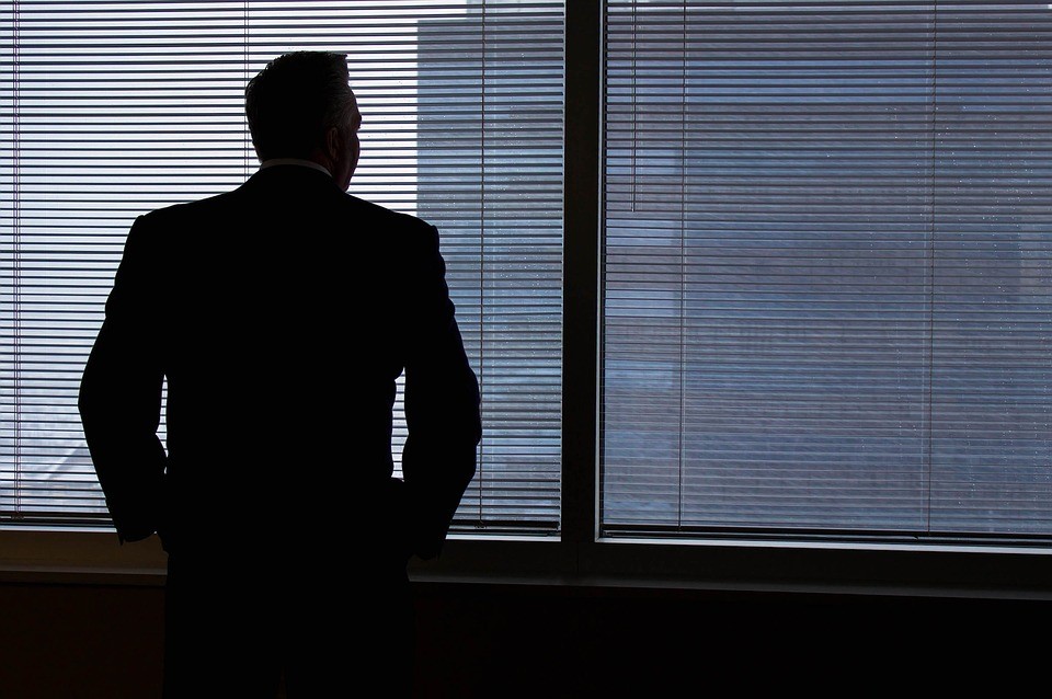 Man in suit staring outside of window with drawn blinds